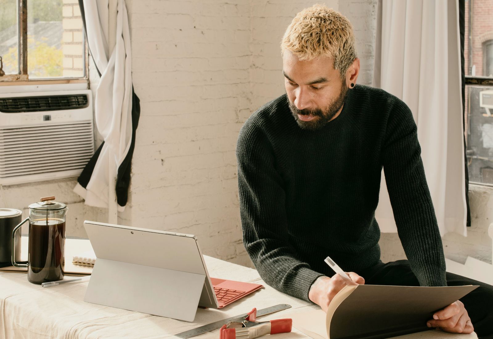 A studio owner in a dark sweater at a linen-covered work table in a sunlit loft, pen in hand, leafing through a portfolio beside his tablet