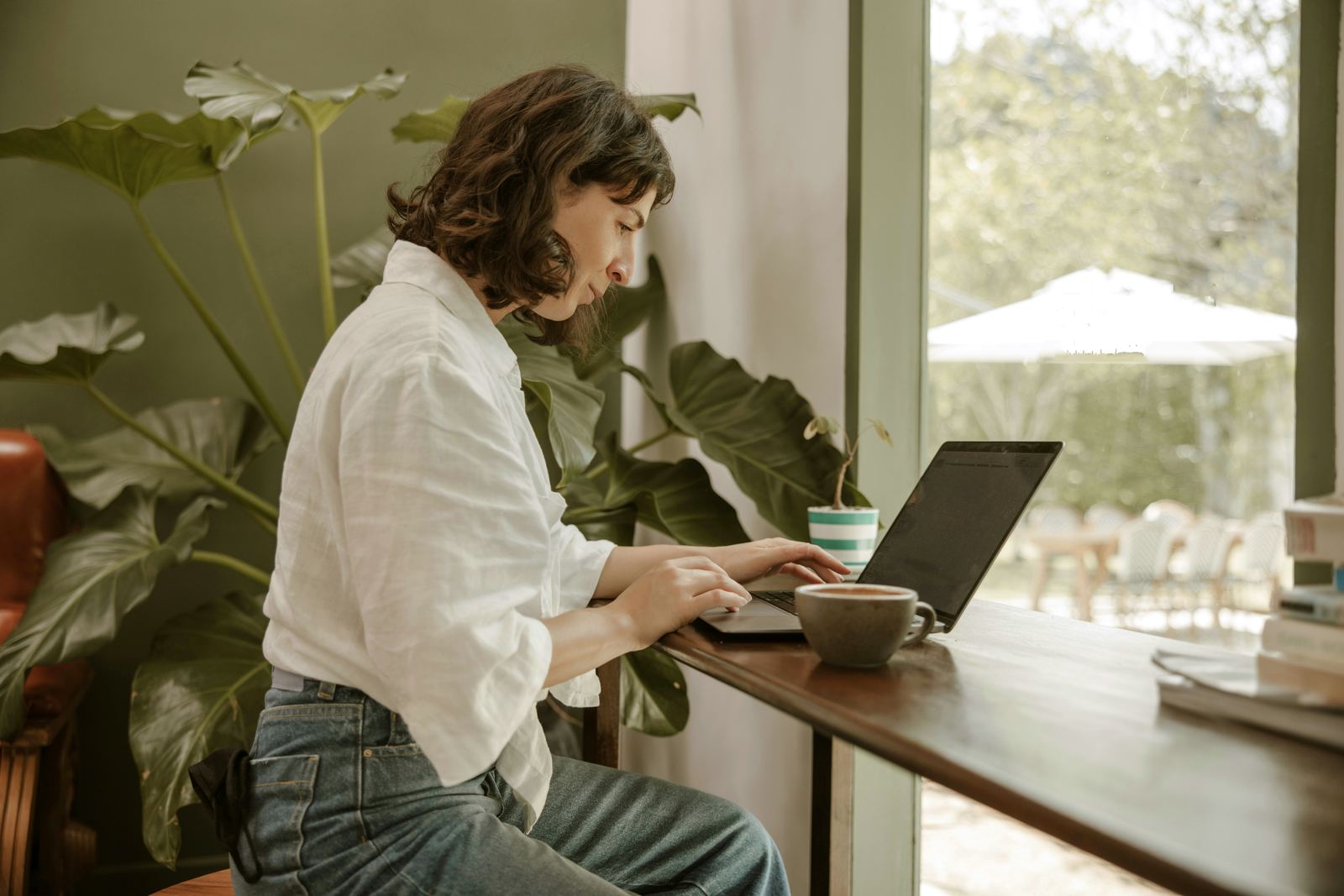 A founder in a linen shirt working on her laptop at a wooden counter by a garden window, beside a large plant